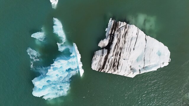 Aerial view of icebergs floating in a glacial lagoon, featuring one large iceberg with distinct dark volcanic ash layers and another smaller, bright blue iceberg in Iceland.