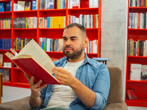 Bearded man wearing a denim shirt sitting in the middle of a bookstore reading a red hardcover book.