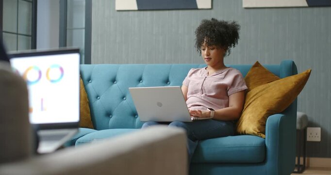 African American woman typing on laptop on teal sofa, glancing at left laptop presence, smiling