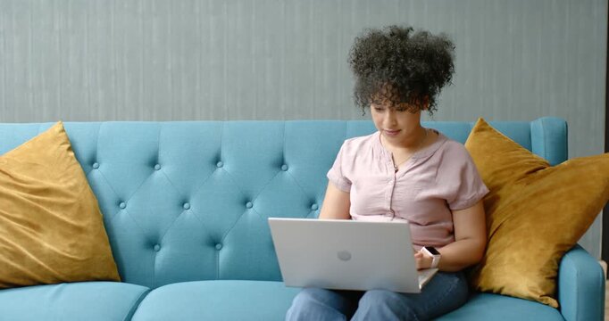 With laptop open, African American woman typing on laptop on teal tufted sofa, studying copy space