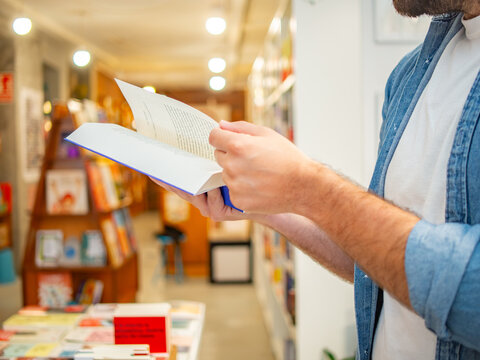 Close-up of an unrecognizable man wearing a denim shirt turning pages of an open blue book in a bookstore or library aisle.