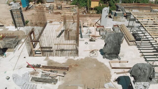 Aerial view over reinforced concrete construction site showing steel rebar cages rising from concrete slab, with stacked timber formwork and materials under bright midday sunlight
