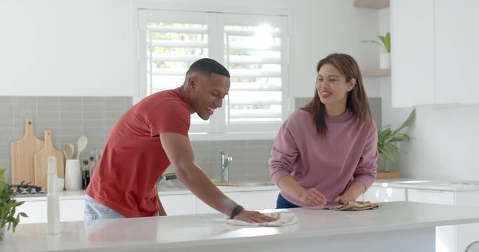 Diverse couple in red tee and mauve top wiping kitchen island, woman grabbing sprayer, teasing man