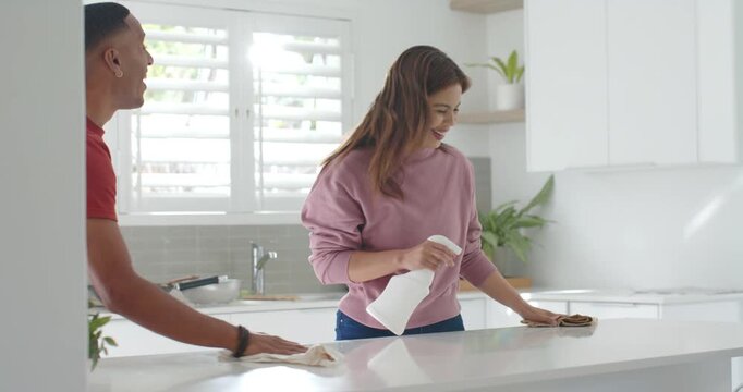 Diverse couple cleaning at white kitchen island, woman miming into spray bottle, man joining wiping
