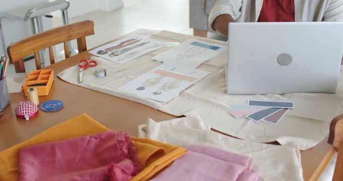 African American man reviewing sketches, aligning fabric swatches at table beside laptop