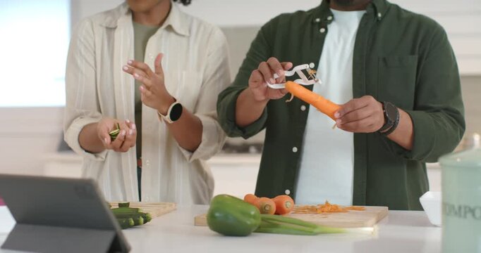 African American couple peeling carrots with peeler in modern kitchen, checking tablet and compost