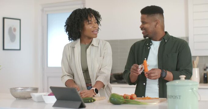 African American couple using tablet recipe chopping veggies peeling carrot and laughing in kitchen
