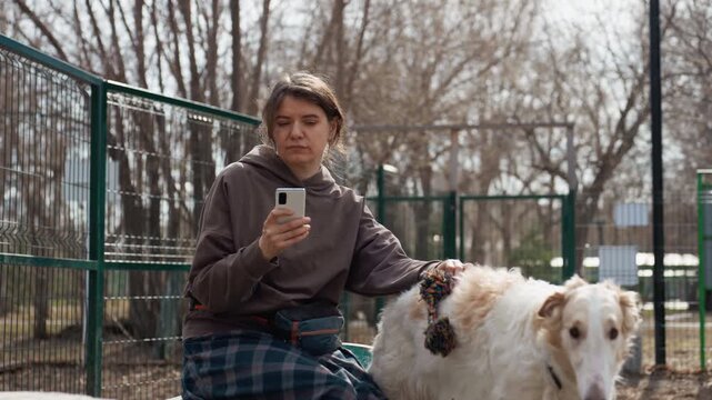 Woman sits on bench in outdoor park holding her smartphone while petting or interacting with large white dog. scene features green metal fencing and leafless trees as background suggesting