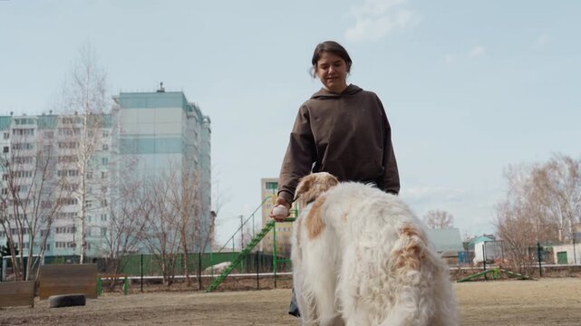 Woman interacts with white and brown dog in outdoor park holding objects for dog to fetch. background features tall apartment buildings bare trees playground equipment like green ladders