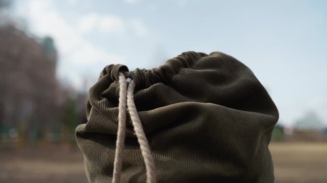Green drawstring bag with visible cord texture swaying gently in outdoor setting. Background shows blurred trees and sky emphasizing fabric detail and natural movement