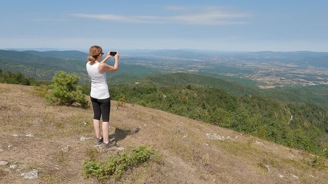 Fit senior woman in top shape and excellent physical condition standing on a mountain top and recording a video of the landscape with a smartphone. Concept of active aging and outdoor hobbies.