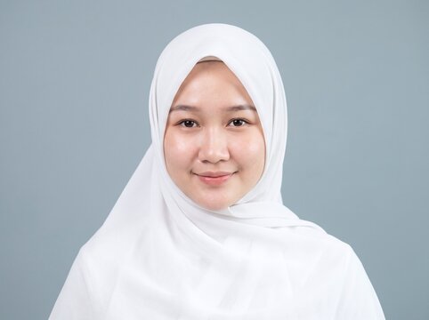 Portrait of a smiling young Muslim woman wearing a traditional white hijab against a simple grey background.