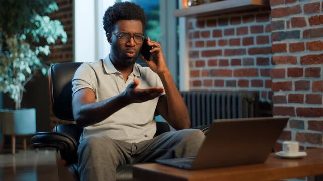 Smiling happy African American business man talking mobile phone call looking at laptop at evening at home office. Cheerful businessman male guy worker agent talk smartphone chatting look at computer