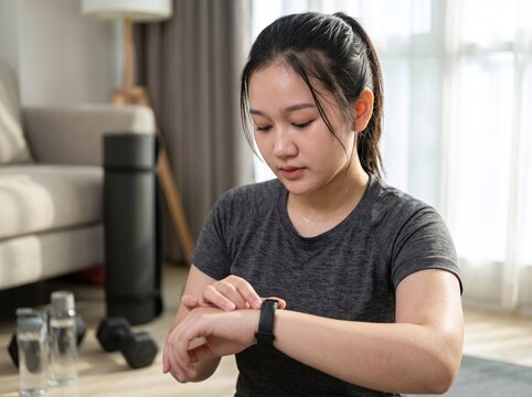 A young Asian woman wearing sportswear looks at her fitness tracker smartwatch while working out in her living room.