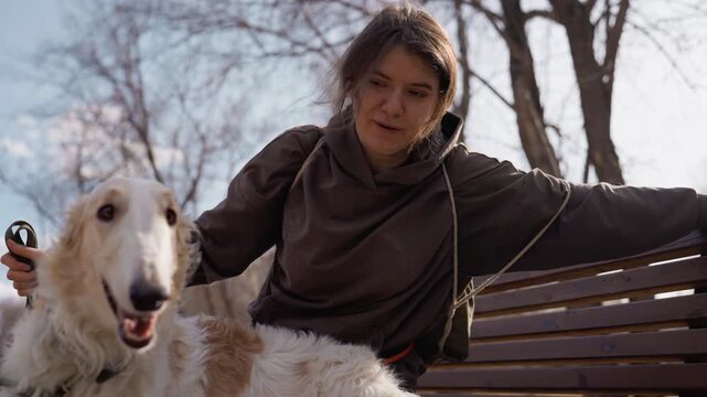 Woman sitting on bench with two large white dogs holding their leashes. woman interacts calmly as dogs appear happy and engaged in outdoor park setting with trees visible in background