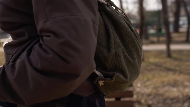 Woman wearing brown hoodie and carrying olive green drawstring bag sits on wooden bench in park. background features bare trees distant buildings other people walking dogs soft natural light