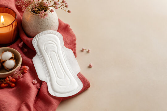 A white, contoured menstrual pad rests on a red cloth next to candles and flowers on a light-colored surface.