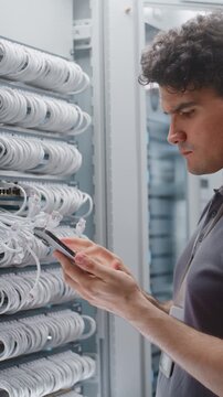 Network Engineer Examines Equipment Connections and Verifies Technical Information on Site, While Female Colleague Maintains Another Cabinet Nearby. Routine Work in Modern Server Facility. Vertical