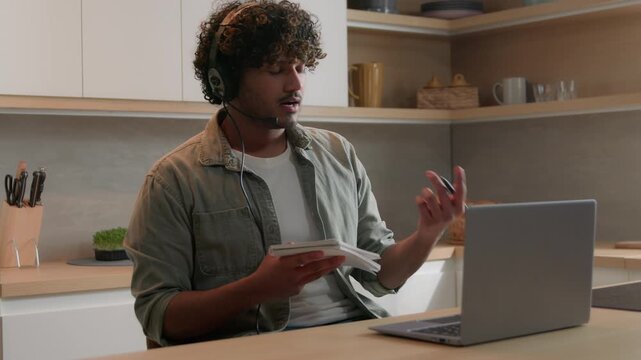 Hispanic Indian Arabian man wearing headphones studying online at home kitchen laptop ethnic male businessman remote video call class elearning student writing notes talking distant lesson education