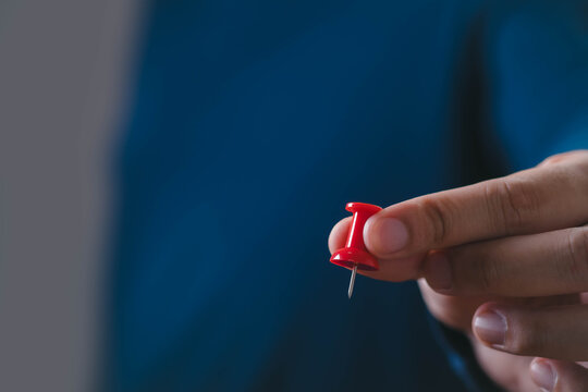 Close-up of a hand holding a red pushpin against a blue background, symbolizing business goals, strategic planning, decision making, target setting, focus, leadership, and project management.