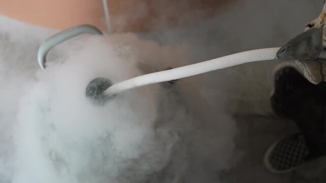 Liquid nitrogen cryogenic fog science experiment laboratory hose releasing vapor cloud near container, cold demonstration on tile floor with gloved hand, dewar safety equipment close view