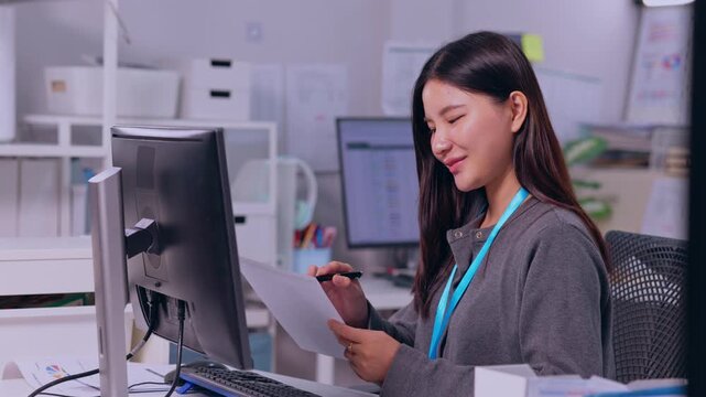 Asian woman office worker and professional employee reviewing document computer monitor while holding smartphone modern workplace, focused business activity with identification lanyard desk during