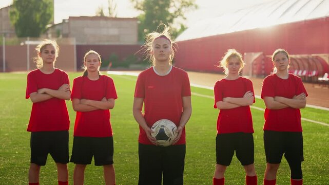 Focused womens soccer team in red training jerseys standing in formation on sunny outdoor field, captain holding ball and preparing for determined practice session