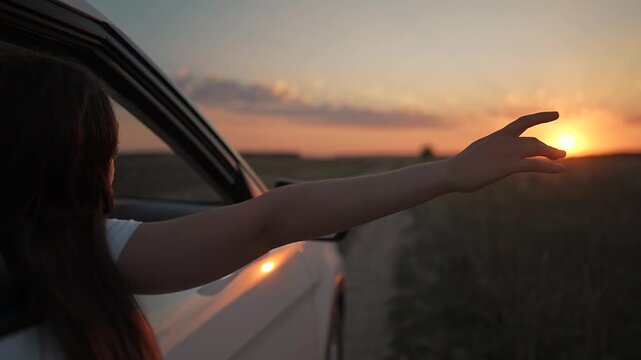 Travel by car through nature road at sunset. Girl feels summer wind on hand. Adventure begins at sunset with car on road. Girl explores summer sunset. Hand of girl moves in wind on travel road