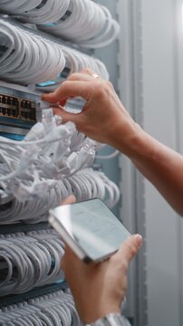 Female Specialist Verifies Patching Against Digital Infrastructure Records on Smartphone, Checking Rack Connections and Port Labels. Concept of Services Equipment in Modern Data Center. Vertical Shot.