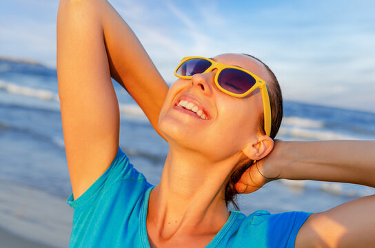 A happy young woman wearing yellow sunglasses smiles while relaxing on a sunny beach near the ocean.