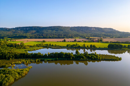 Aerial view of Zabakor pond surrounded by lush green fields, dense forests, and rolling hills under a clear blue sky in Brezina, Central Bohemian Region, Czechia.