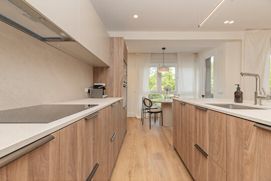 Modern galley kitchen with wood cabinetry, white countertops, and a bright dining nook framed by large windows.
