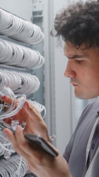 Male Technician Verifies Ethernet Patching, Physical Port Labels Against Network Documentation on smartphone. Female Colleague Performs Maintenance on Adjacent Server Rack in Facility. Vertical Shot.