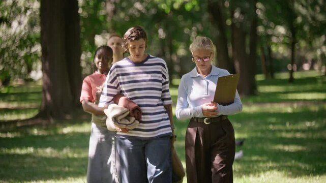 Diverse multiethnic group walking outdoors in park carrying chairs and mats with Caucasian senior psychologist holding clipboard. Clip illustrates outdoor psychotherapy or mental wellness retreat