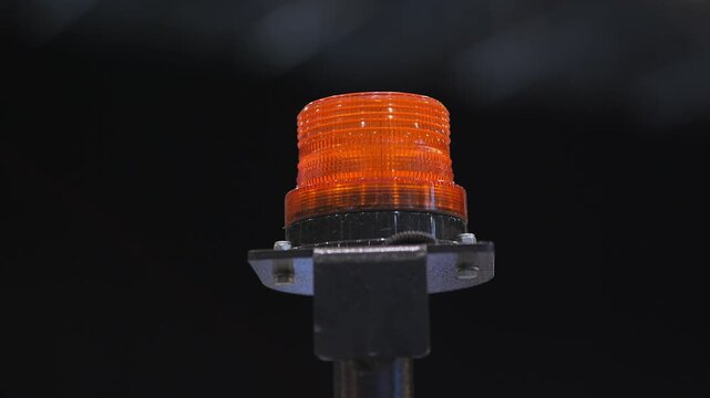 Bright orange warning light shines steadily against a dark background. The light is mounted on a metal pole. Its glow is intense and focused. Small particles float near the light source