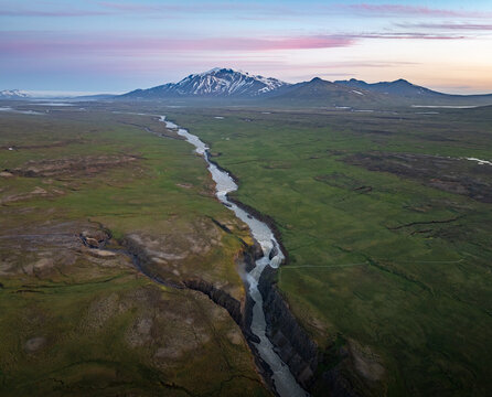 Aerial view of a winding river flowing through a deep canyon in the vast green landscape, with snow-capped mountains in the background under a soft twilight sky Iceland.