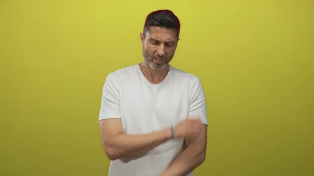 Man rubbing his upper arm and wincing while wearing a white t shirt against yellow studio wall; discomfort fatigue.