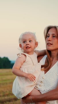 Smiling young mother holding blonde toddler daughter in white summer dresses while enjoying a peaceful sunset moment together in an open countryside field