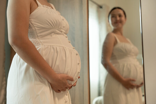 Close-up of a pregnant woman's belly in a white button-front dress, her smiling reflection visible in the mirror behind.