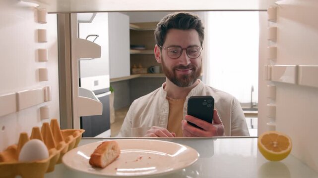 Happy Caucasian hungry man wearing glasses opening empty refrigerator ordering food delivery with mobile app POV inside fridge male guy buying groceries online supermarket cellphone service indoors