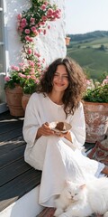 Obraz premium Woman in white dress eating cake on flower terrace with white cat and vineyard view for relaxed lifestyle banner and cottagecore summer background