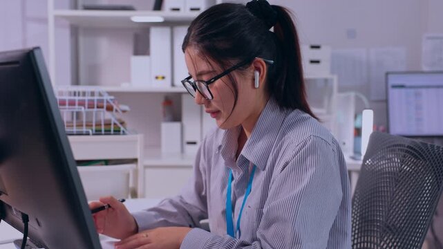Asian woman office worker and female employee wearing eyeglasses and wireless earbuds, analyzing computer monitor while holding pen during focused desk work at modern workplace desk setup