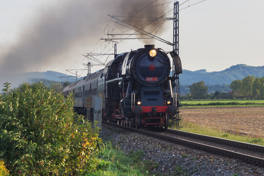 View of the Albatros 498 104 steam locomotive pulling passenger cars through the rural landscape with mountains in the background near Velka Luka, Banska Bystrica Regio, Slovakia.