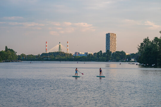 View of two people paddleboarding on the calm waters of An der Oberen Alten Donau with a distant modern skyline and cable-stayed bridge in Wien, Wien, Austria.