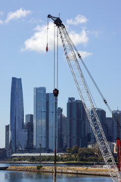 View of a lattice boom crane with a heavy hook against the modern high-rise skyline of Barangaroo, New South Wales, Australia.