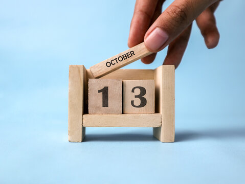 A woman's hand holds a wooden block with October written on it on a wooden block calendar for October 13th against a light blue background.