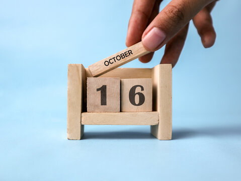 A woman's hand holds a wooden block with October written on it on a wooden block calendar for October 16th against a light blue background.
