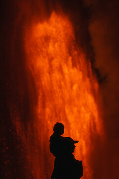 View of a silhouette of a person carrying a child on their shoulders against the backdrop of a glowing lava fountain at Kilauea Crater in Hawaii County, Hawaii, United States.