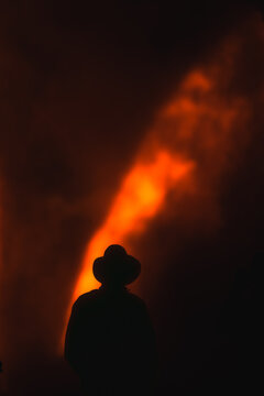 View of a person in a hat silhouetted against the glowing lava plume and volcanic gases rising from Kilauea Crater at night in Hawaii County, Hawaii, United States.
