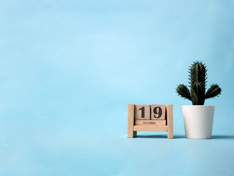 A wooden block calendar for October 19th with a plastic cactus plant on the side on a light blue background.